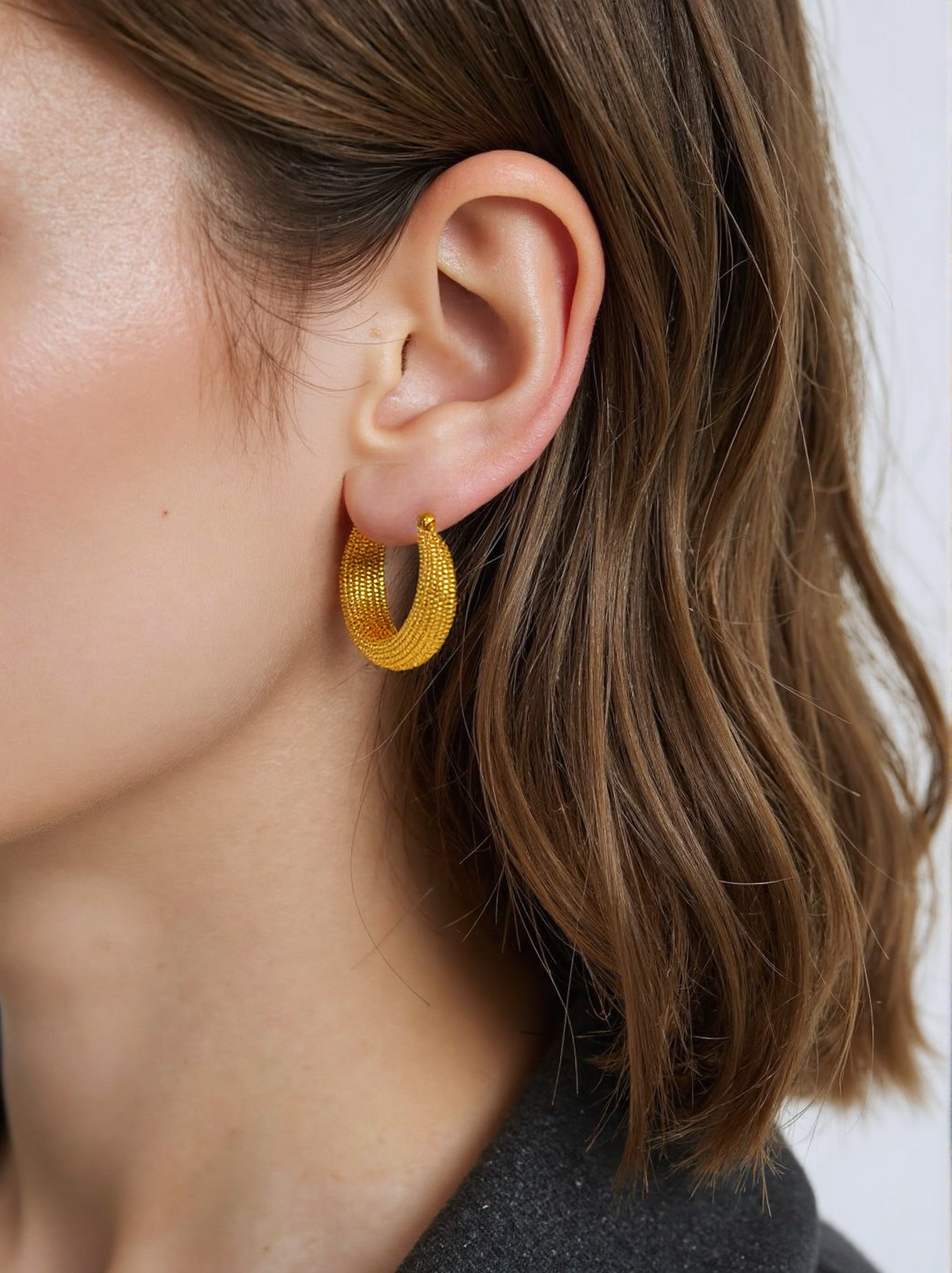 A woman wearing gold textured hoop earrings, featuring an intricate design with a polished finish. These statement hoops add a bold yet elegant touch to her outfit.