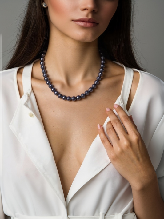 A woman wearing an elegant black pearl necklace with round simulated pearls, styled with a white satin blouse and a gold ring.