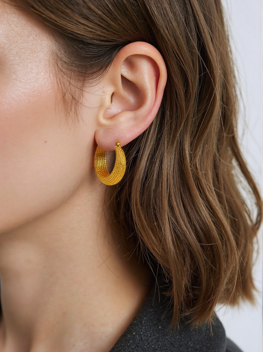A woman wearing gold textured hoop earrings, featuring an intricate design with a polished finish. These statement hoops add a bold yet elegant touch to her outfit.