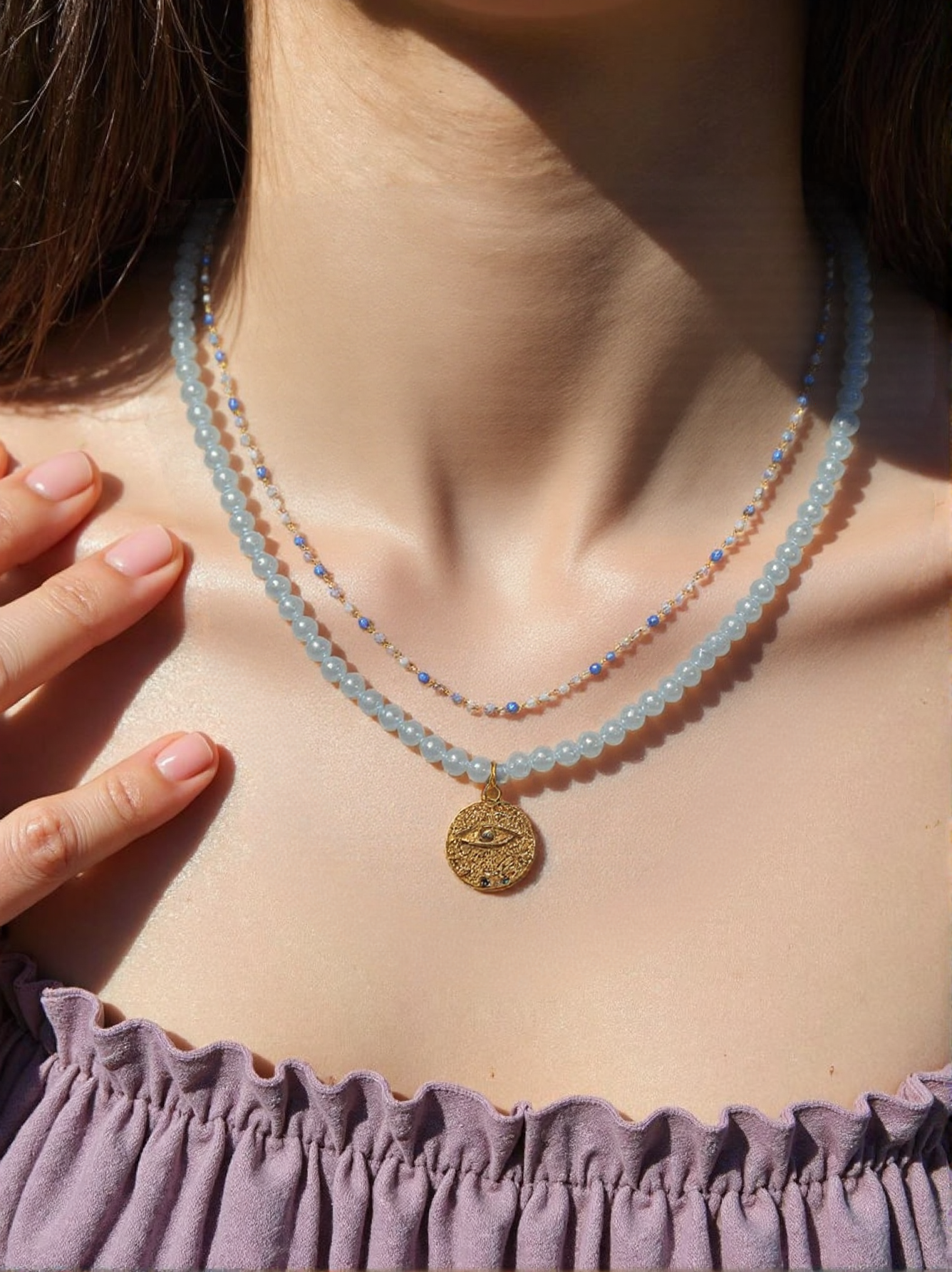 A woman wearing a layered blue beaded necklace with a gold evil eye pendant, styled with a bohemian off-shoulder outfit under natural sunlight.